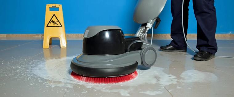 Bodenreinigungsmaschine mit roter Bürste auf nassem Fliesenboden, dahinter ein Warnschild